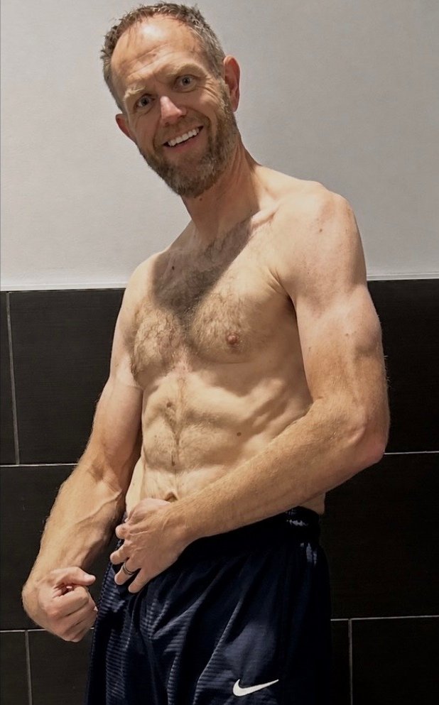 Lean man posing shirtless against black tiled wall after transformation