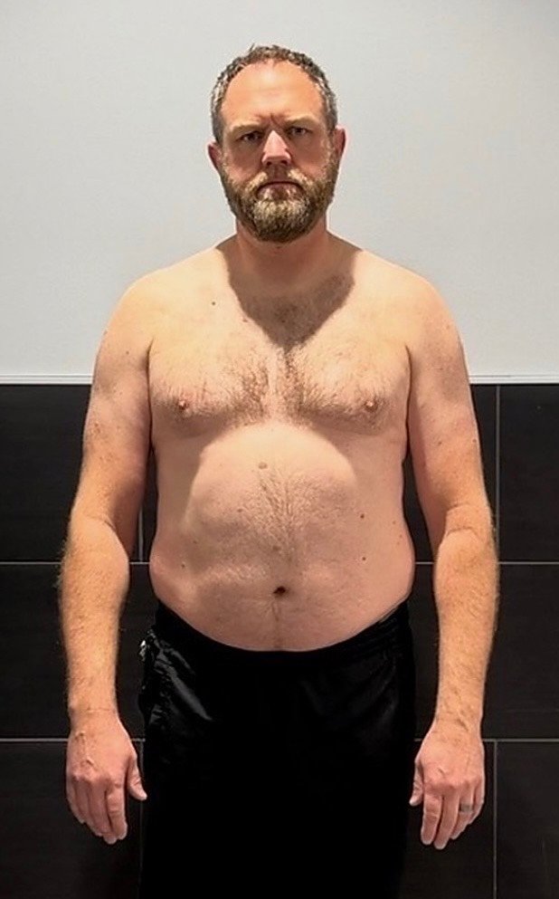 Man standing shirtless against black tiled wall before fitness transformation