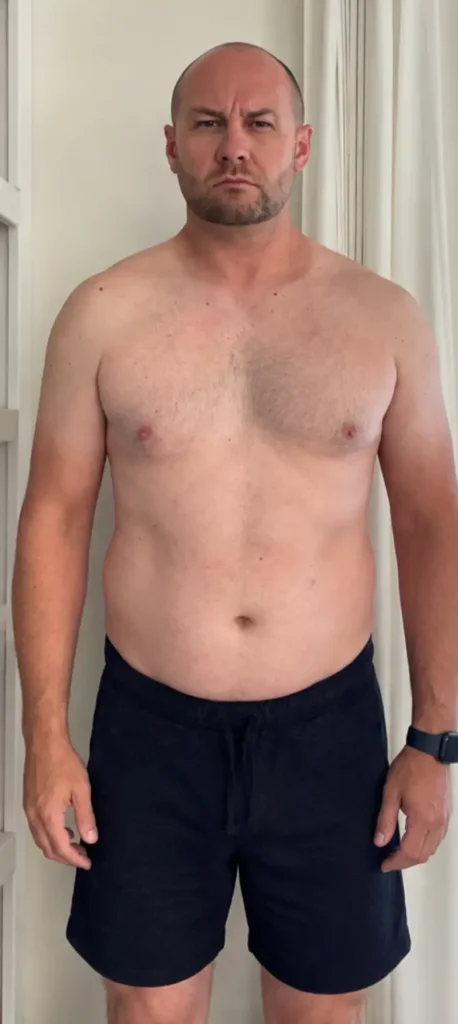Bald man standing shirtless indoors before fitness transformation