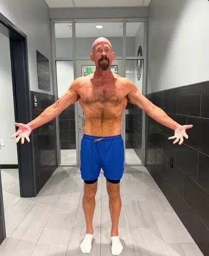 Older muscular man standing shirtless with arms extended indoors